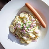 Creamy potato salad with sausages, garnished with chives, a flavorful German classic.