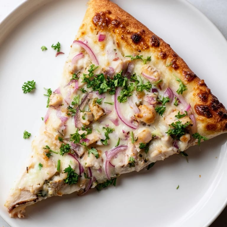 A slice of Chicken Alfredo Pizza is lifted, revealing creamy sauce, tender chicken, and a crispy edge.